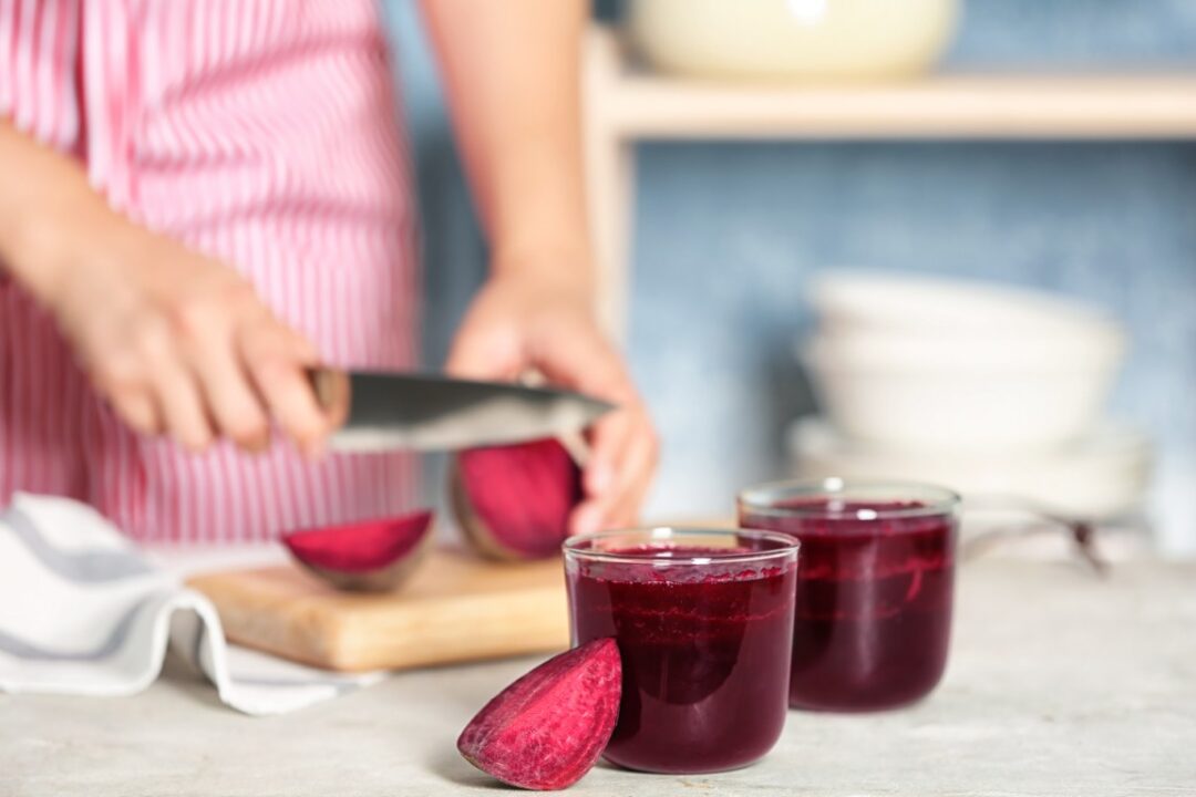 2 Gläser mit Rote Bete-Saft, im hintergrund eine verschwommene Perosn, die Rote Bete schneidet.